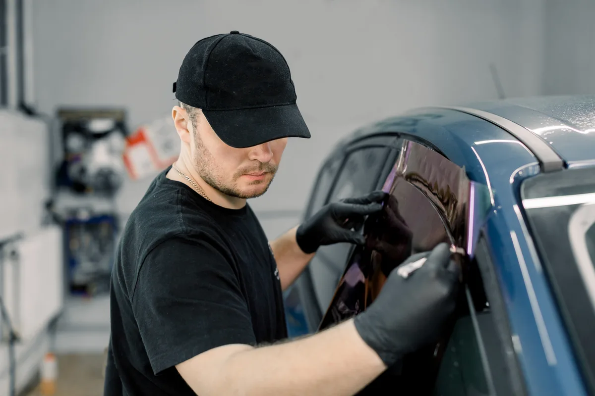 A window tint installer smoothing ceramic film onto a car door window inside a clean, well-lit shop bay
