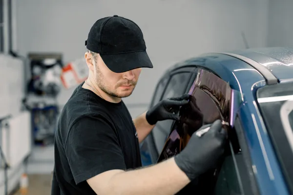 A window tint installer smoothing ceramic film onto a car door window inside a clean, well-lit shop bay