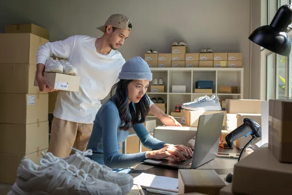 An online store owner packing customer orders at a laptop surrounded by shipping boxes