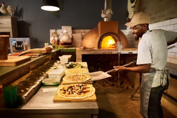 A pizzeria cook sliding a fresh pie into a wood-fired oven during a busy dinner service