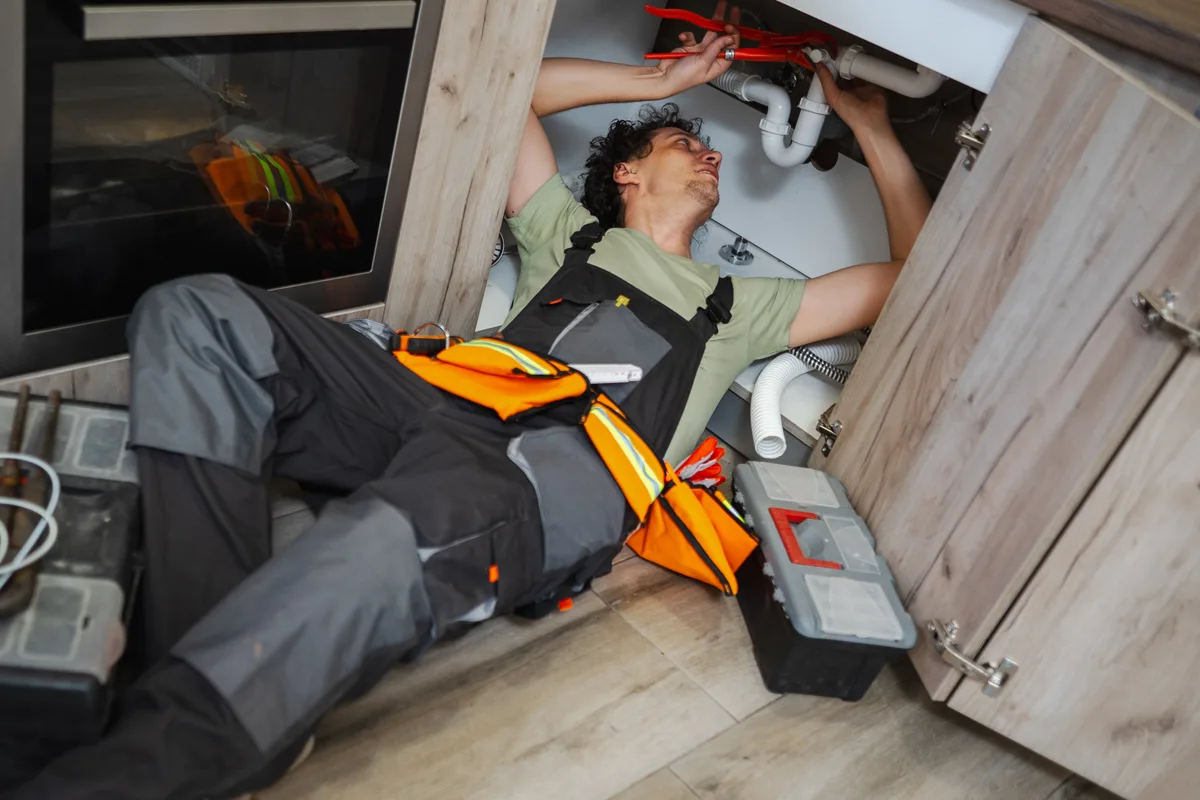 A uniformed plumber repairing a pipe under a kitchen sink, representing a booked residential service call