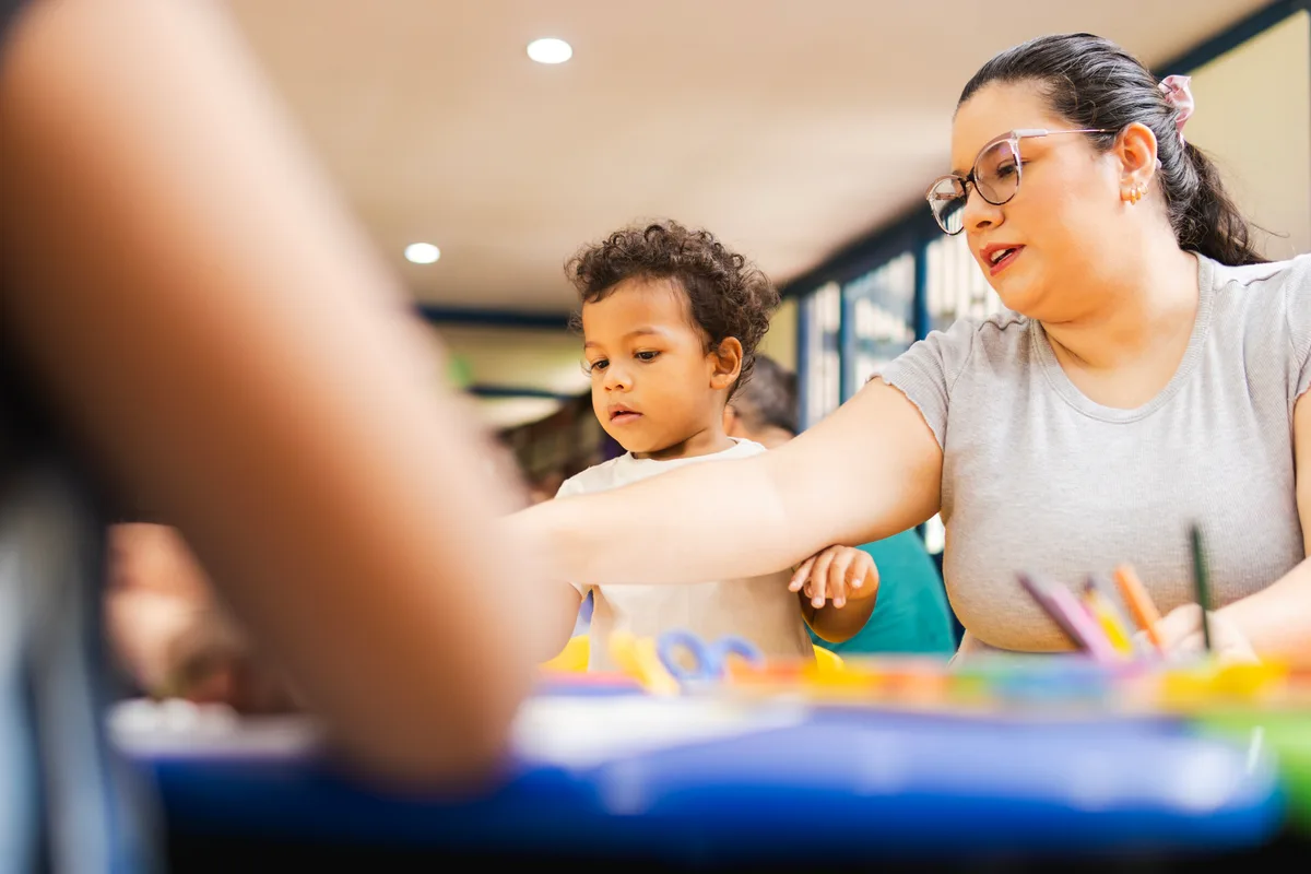 A preschool teacher sitting with a small group of toddlers at a low table in a bright, modern daycare classroom