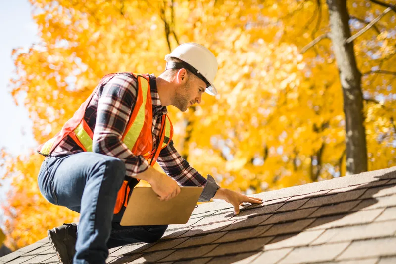 Beacon's lead estimator in a hard hat and safety vest kneeling on a shingle roof with a clipboard, autumn trees behind