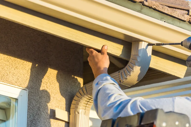 An installer fastening a new white aluminum gutter and downspout elbow to the fascia under an eave