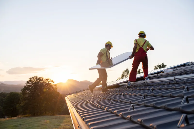 Two Beacon roofers in red coveralls and hard hats carrying a panel up a dark standing-seam roof at sunset, with wooded hills behind them