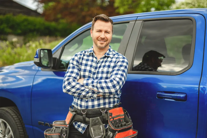 Beacon's owner in a plaid shirt and tool belt, arms crossed, standing beside a blue pickup