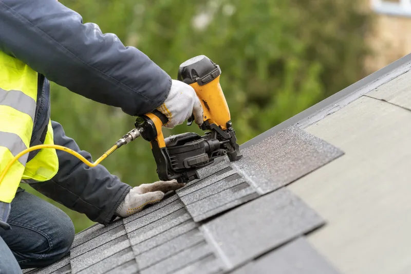 Close view of a roofer in gloves and a safety vest nailing a replacement shingle with a coil nailer