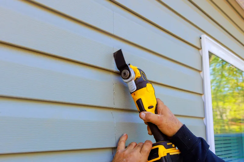 Gloved hands using an oscillating tool to cut out a damaged run of pale blue lap siding
