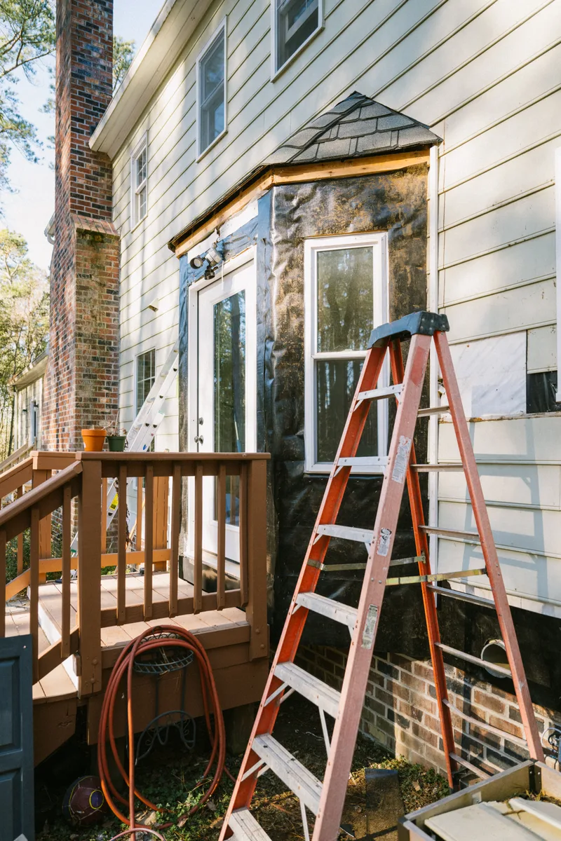 Back corner of a two-storey house with grey lap siding, a wooden deck and an orange ladder set up for siding work