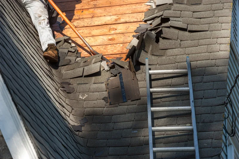 A roofer tearing worn asphalt shingles off down to the wood deck, with a ladder resting on the roof