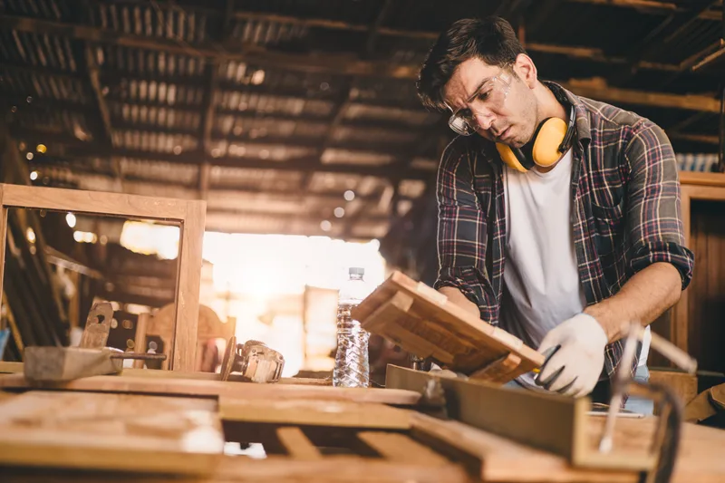 A carpenter in safety glasses and ear defenders shaping a piece of timber at a workbench in a sunlit wood shop