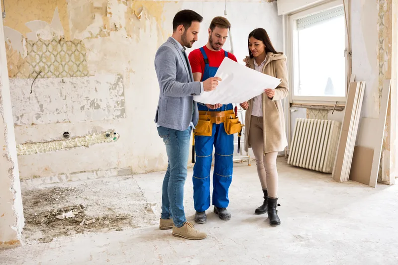 A contractor in work overalls and a tool belt reviews a floor plan with two homeowners inside a gutted room