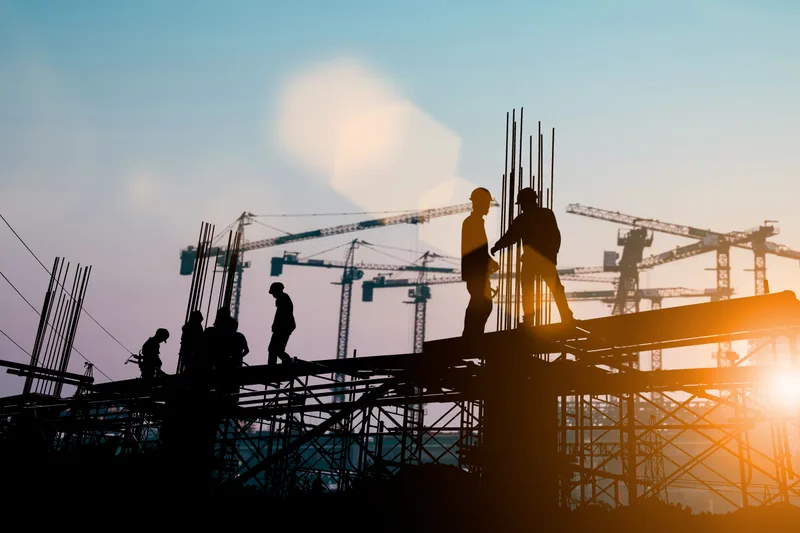 Construction crew silhouetted on a steel frame at sunrise, with tower cranes behind