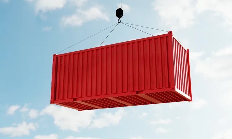 Red shipping container suspended mid-air from a crane hook against a blue sky