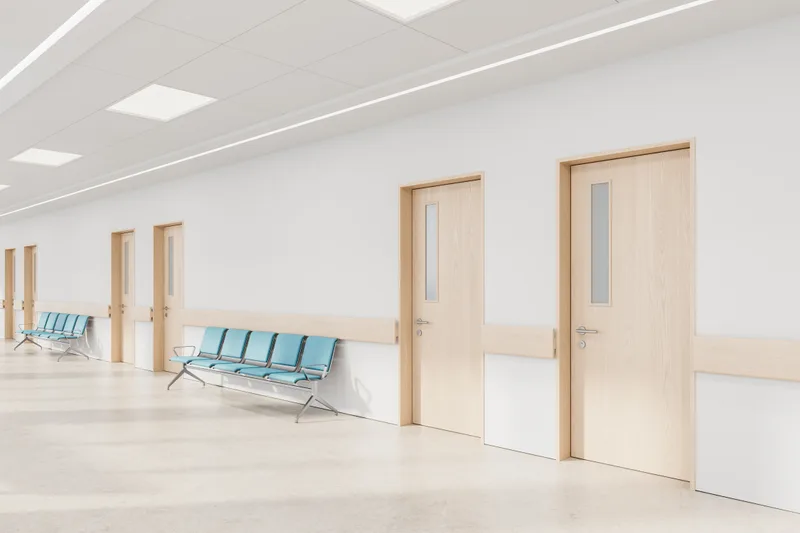 Bright clinical corridor with pale wood doors, blue waiting chairs and a polished floor