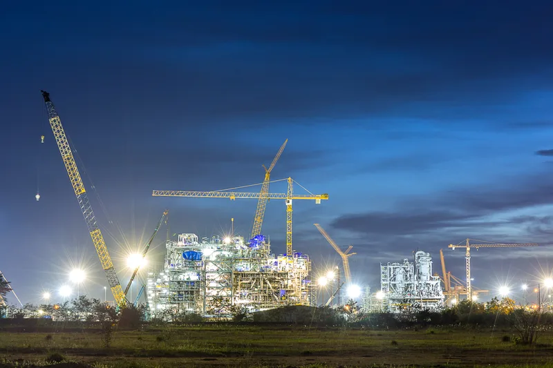 Process plant under construction at night, lit by site lights, with crawler cranes against a deep blue sky