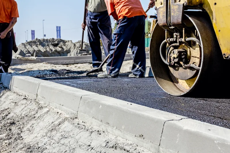 Crew finishing new asphalt against a fresh concrete curb as a roller passes