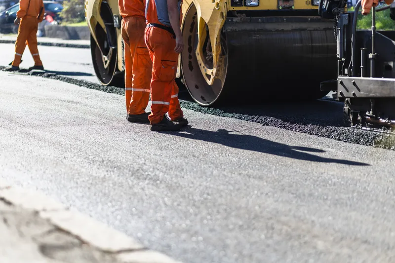 Roller compacting a new asphalt lane while two crew members check the edge