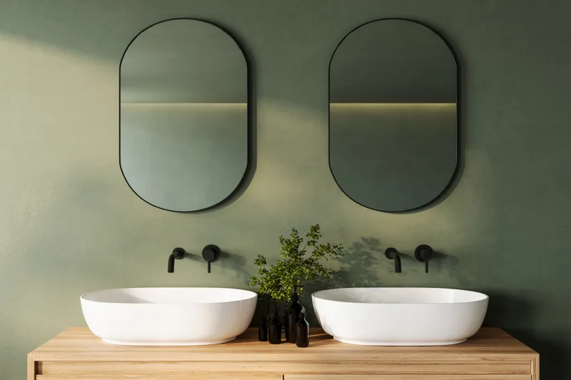 Bathroom with a sage green plaster wall, two pill-shaped mirrors, matte black wall faucets, and twin white vessel sinks on an oak vanity