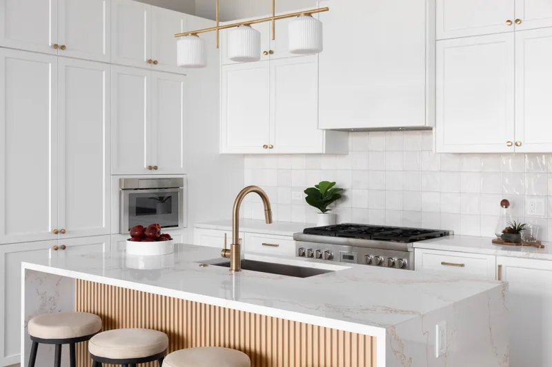Remodeled white kitchen with a marble waterfall island wrapped in vertical oak slats, brass faucet, and three counter stools
