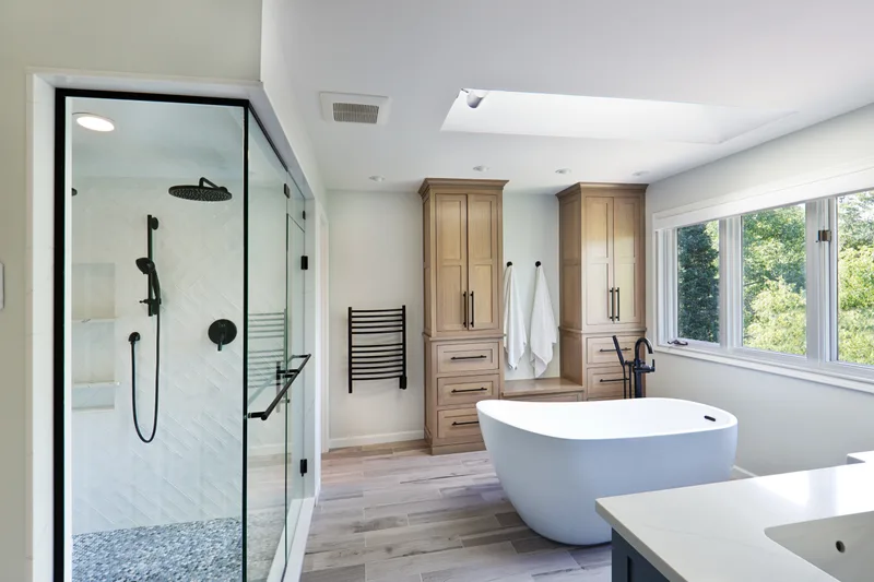 Bright primary bath with a walk-in glass shower in a slim black frame, a freestanding tub under a window, and oak storage towers