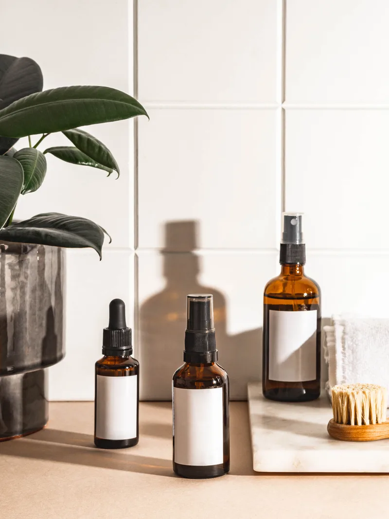 Amber glass bottles with plain white labels on a tiled bathroom shelf, a green plant and a wooden brush beside them in soft morning light