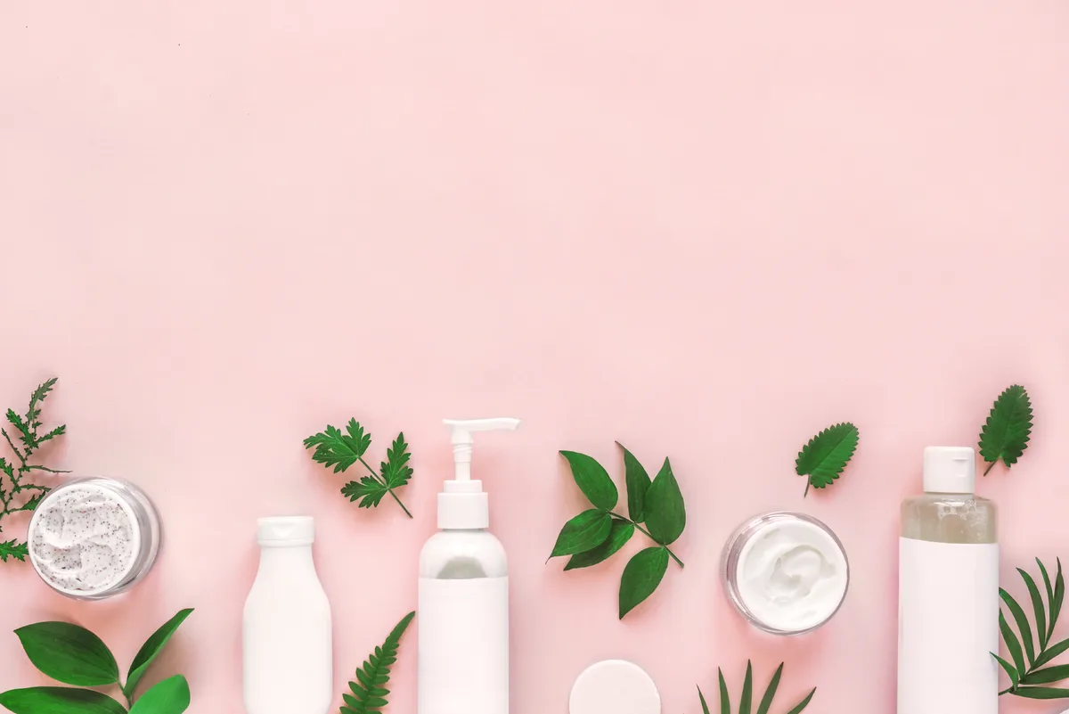 White skincare bottles, jars, and a pump dispenser arranged in a row with fresh green leaves on a blush-pink surface