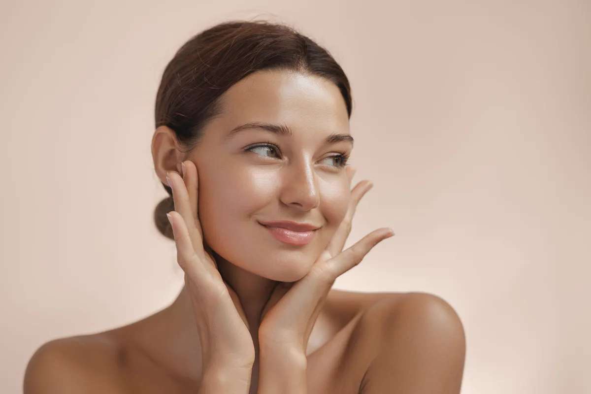 Woman with calm, clear skin resting her fingertips on her cheeks against a warm beige backdrop