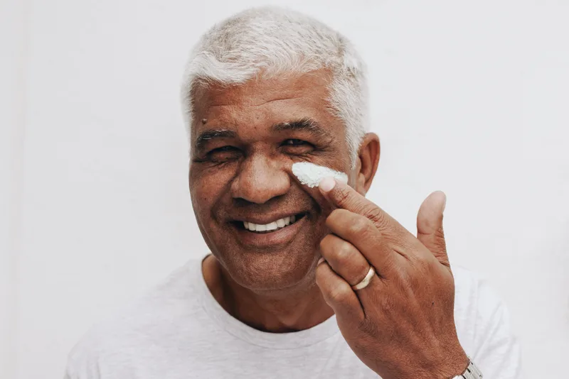 A smiling older man dabbing white moisturizer under his eye