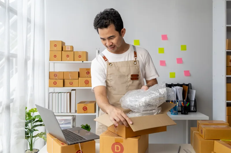 A team member in an apron packing an order into a cardboard box in a bright stockroom