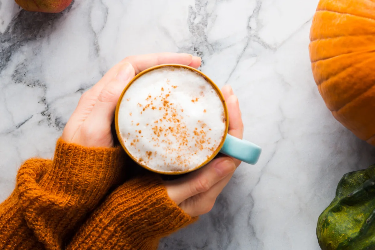 Hands in a warm orange sweater cradling a cinnamon-dusted latte on a marble table beside a pumpkin