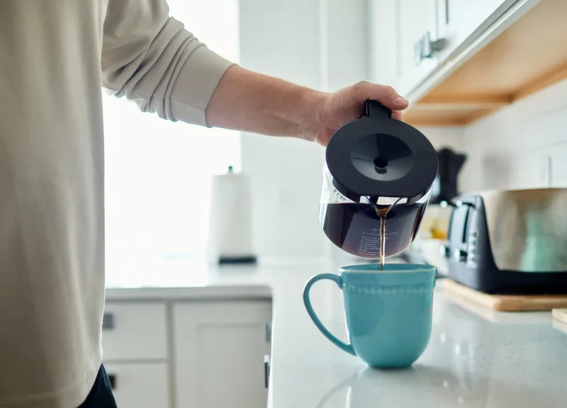 Person pouring fresh coffee from a carafe into a blue mug in a bright kitchen
