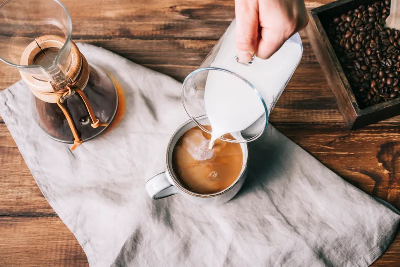 Milk poured into a mug of fresh coffee on a wooden table, a glass pour-over carafe and a tray of beans beside it