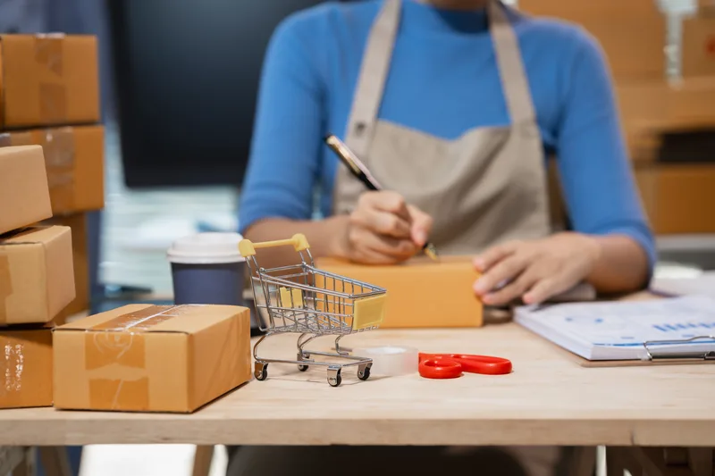 A packer in a canvas apron labelling a kraft shipping box at a bench stacked with orders