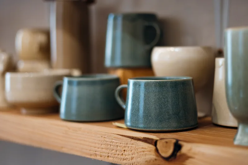Handmade stoneware mugs in sea-glass blue and oat cream glazes on a wooden shelf