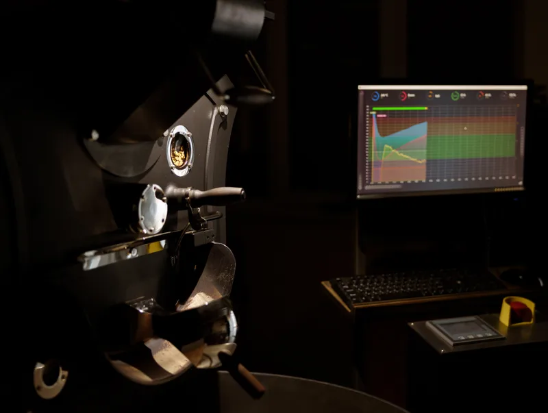 A drum coffee roaster in a dim room beside a monitor tracking the roast curve