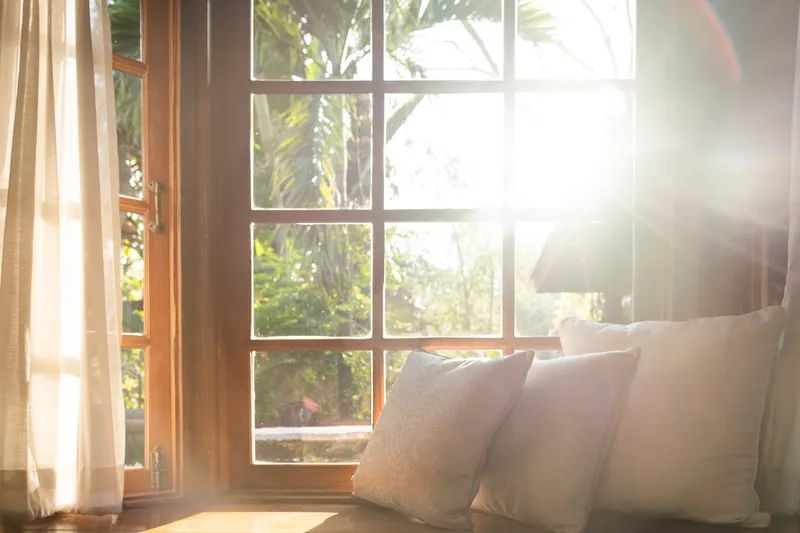 Three washed-linen cushions arranged on a wooden window seat in warm morning light, palm leaves visible through the panes