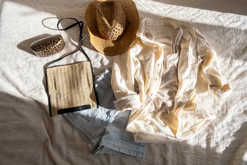 Flat lay of an ecru linen shirt, denim shorts, straw hat, and raffia bag in warm sunlight on a textured bedspread