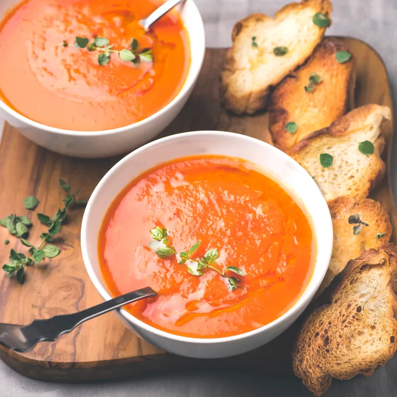 Two bowls of tomato-thyme soup with torn golden croutons on an olive-wood board