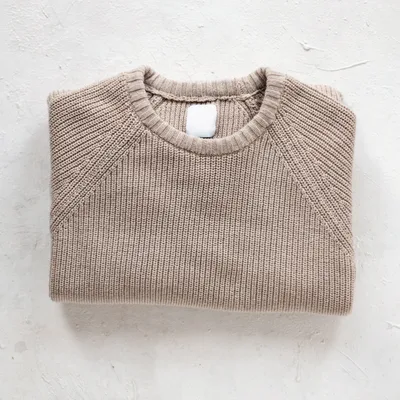 Oat-coloured fisherman-rib cotton crewneck sweater, folded on a white plaster surface