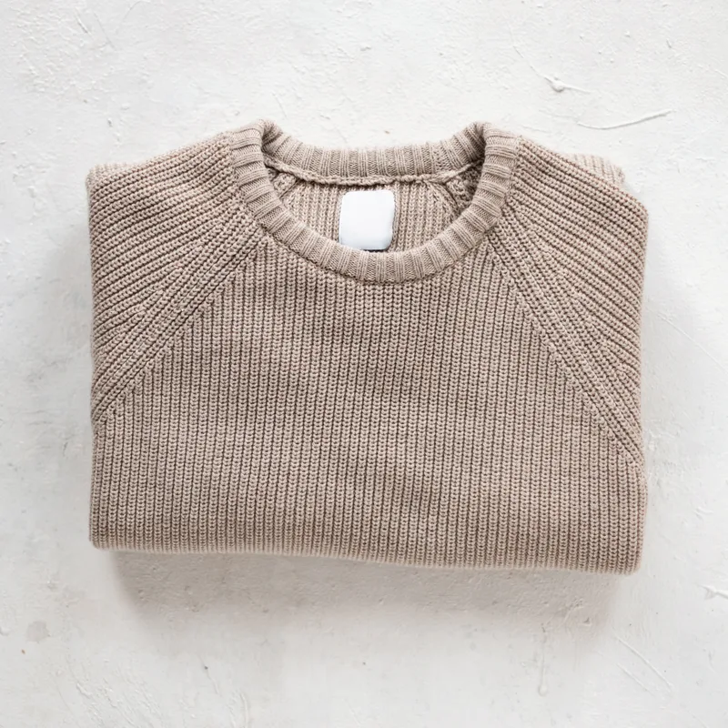 Oat-coloured fisherman-rib cotton crewneck sweater, folded on a white plaster surface