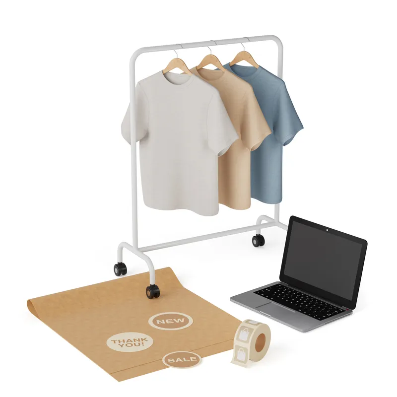 Packing bench with garment-dyed tees on a rolling rack, kraft wrapping paper, thank-you stickers, and an open laptop