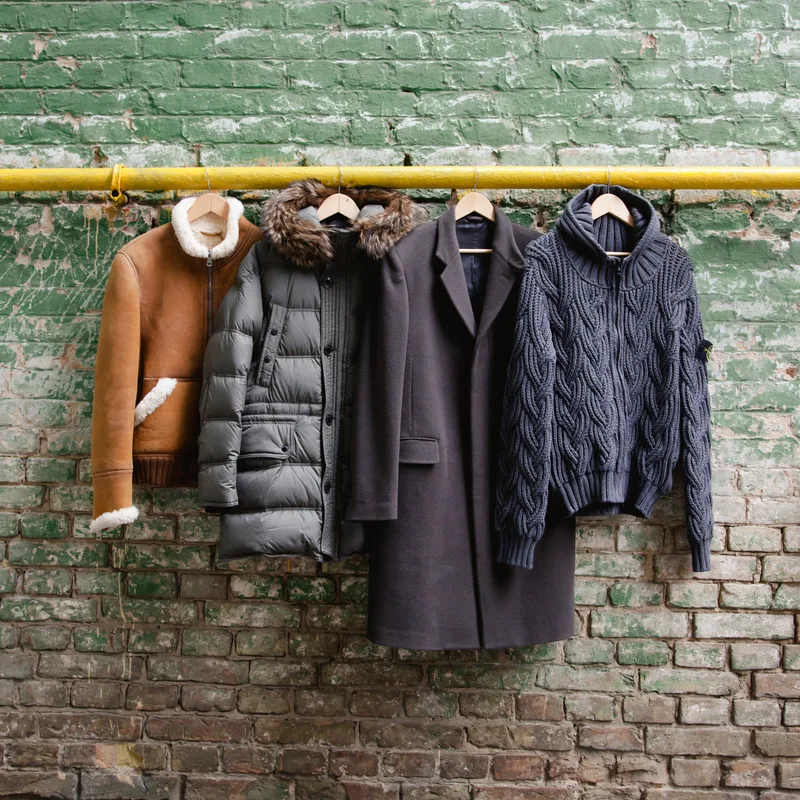 Four winter layers hanging from a yellow rail against a green brick wall: shearling jacket, quilted parka, wool overcoat, and cable-knit zip-up