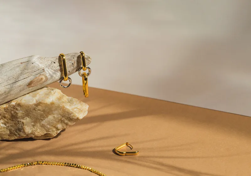 Gold link earrings hanging from driftwood above a sandstone, with a fine gold chain on a warm beige ground