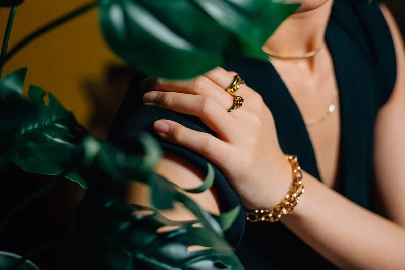 Model resting a hand on her shoulder wearing the sculptural gold Aurea signet rings, styled with a black dress and monstera leaves