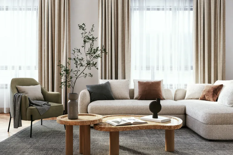 Sunlit living room with a cream boucle sectional, an olive-green armchair, and two nested oak coffee tables on a grey wool rug
