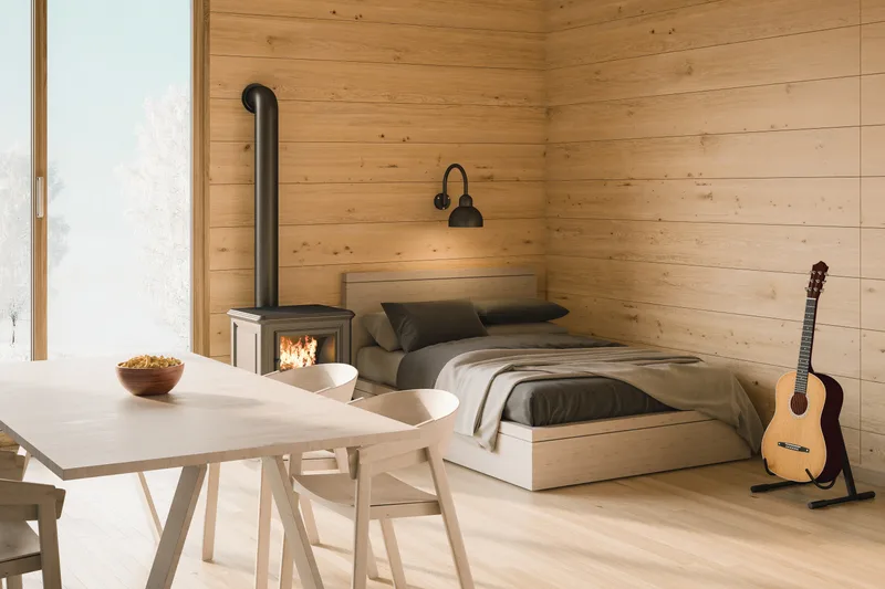 Pine-panelled room with a light-oak platform bed, a pale trestle dining table with two chairs, and a black wood stove