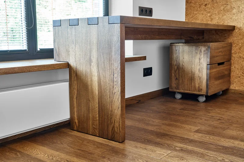 Close view of a solid oak desk with box-jointed corners and a matching oak drawer unit on castors, beside a sunny window