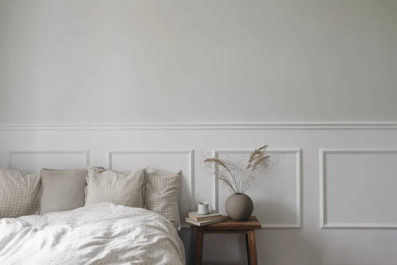 Calm bedroom with rumpled white linen, a small wooden stool used as a nightstand and a vase of dried grass against a panelled wall