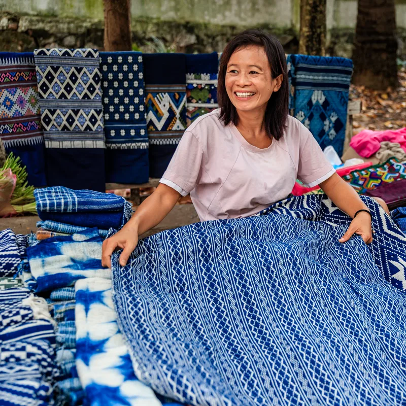 A textile maker smiling as she unfolds a length of indigo-dyed patterned cloth at an open-air workshop, woven fabrics hanging behind her